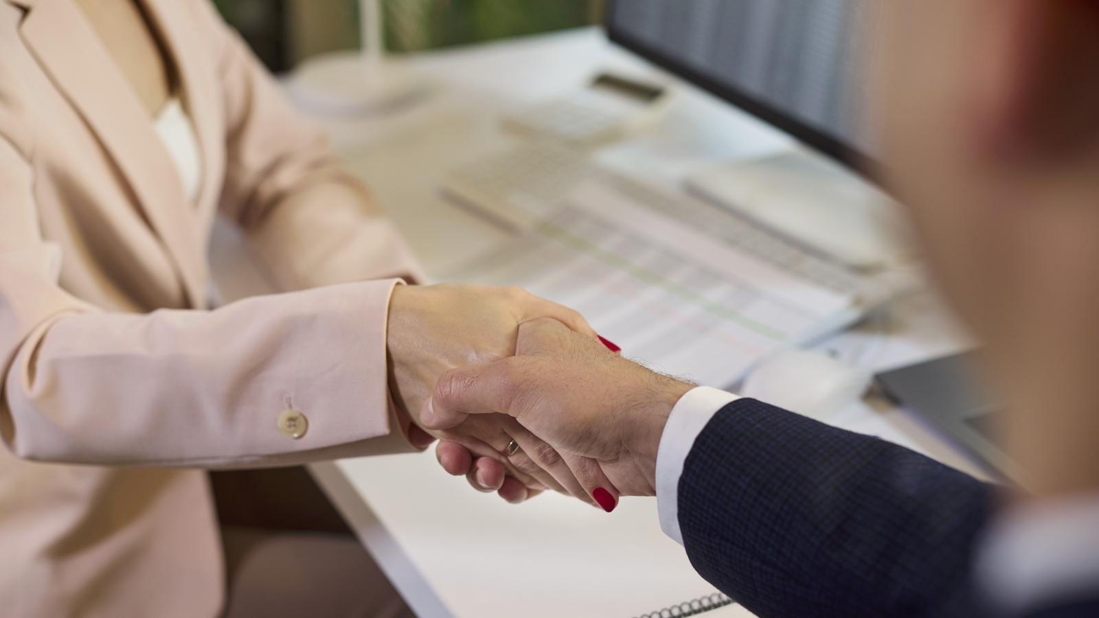 A closeup of two businesspeople shaking hands while sitting in front of a computer.
